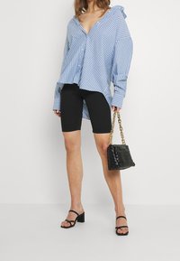 Nadměrná košile s modrobílými pruhy na knoflíky, černé cyklistické šortky a černé sandály na blokovém podpatku s pásky. Drží černou pletenou kabelku.