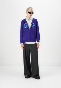 Persoon in een paarse GAP-hoodie, gestreepte overhemd, wijde donkere broek, zwarte chunky schoenen en zwarte zonnebril staande op een witte achtergrond.