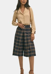 Blusa beige con bottoni abbinata a pantaloni culotte a vita alta a scacchi blu e marrone, con pieghe e dettaglio della cintura. Tessuto con una texture liscia.