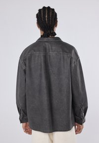 Giacca di jeans nera, oversize, con una texture cerata, polsini con bottoni e dettaglio alla schiena, caratterizzata da una vestibilità rilassata e un design minimalista.