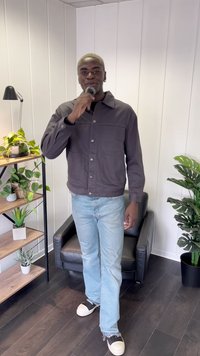 Giubbotto grigio scuro con bottoni, dotato di due tasche frontali, abbinato a jeans azzurri chiari e sneakers nere. Ambientazione interna con piante e mobili.