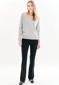 Lichtgrijze v-halstrui van zachte stof, gecombineerd met strakke zwarte broek. Zwarte glanzende schoenen maken de outfit compleet tegen een witte achtergrond.