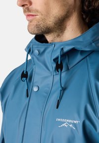 Blå vattentät jacka med hög krage, justerbara snoddar, svarta detaljer och vit logotyp. Slät textur och praktisk design.