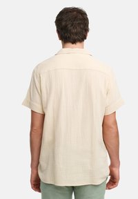Camisa de manga corta color crema con una tela texturizada, que presenta un cuello puntiagudo y un ajuste relajado, vista desde atrás.
