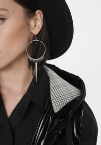 Duża srebrna kolczyk w kształcie obręczy z czarnymi frędzlami i spiczastym zawieszką. Osoba nosząca ma ciemne włosy, czarną kurtkę i szary kołnierzyk w kratkę.