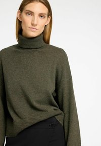 Olivgrüner, grob gestrickter Pullover mit hohem Rollkragen, überschnittenen Schultern und geripptem Saum. Kombiniert mit schwarzer, taillierter Hose mit Knopfdetails.