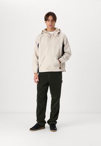 Beige pullover jacket with a hood, side navy panels, snap buttons, large front pocket, paired with dark green corduroy pants and black sneakers.