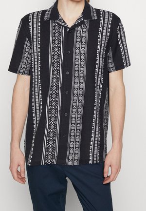 Schwarzes kurzärmeliges Hemd mit weißen vertikalen Stammesmustern, getragen von einer stehenden Person mit marineblauen Hosen.