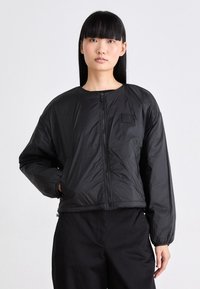 Vrouw met steil zwart haar, gekleed in een zwarte ritssluiting jas en zwarte broek, staand tegen een effen lichte achtergrond.