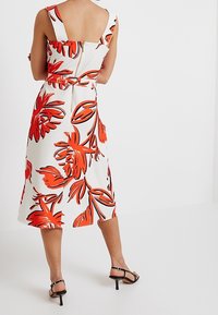 Robe midi florale avec fond blanc, grands motifs de fleurs rouges et noires, corsage ajusté, dos nu et taille nouée. Sandales à talons noires.