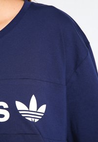 Tmavě modrá mikina s ribbovaným límcem, volným střihem a bílým logem Adidas s trefoil designem na přední straně.