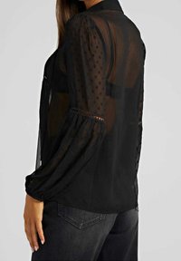 Blouse noire transparente à motif de pois, manches longues bouffantes et détails en dentelle aux poignets. Portée avec un jean foncé.