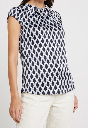 Femme portant un chemisier à manches courtes à motifs géométriques noir et blanc avec un pantalon blanc, debout devant un fond uni.