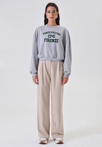 Giovane donna in felpa grigia con il testo "Ponte Vecchio 1345 Firenze", pantaloni beige a gamba larga e sneakers bianche, in piedi su uno sfondo bianco.