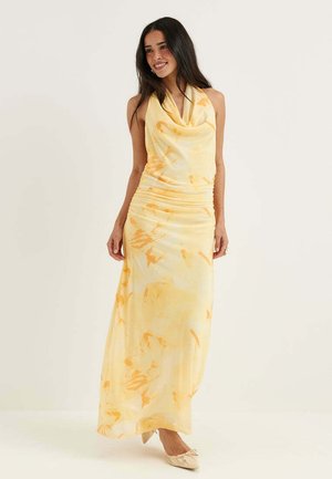 Giovane donna con lunghi capelli scuri che indossa un vestito lungo senza maniche giallo e arancione e ballerine beige, sorridente e in piedi su uno sfondo semplice.