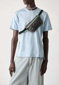 T-shirt en coton bleu clair associé à un pantalon en denim clair. Une banane noire à motifs avec des accents turquoise est portée en travers de la poitrine.