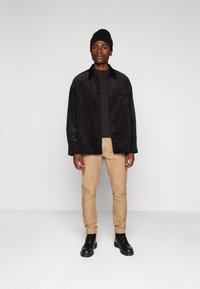 Fekete kordbársony gombos ing egy fekete póló fölött, bézs chinos nadrág és fekete fűzős csizma. Egyszínű darabok, látható minták nélkül.