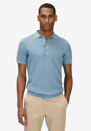 Man standing with hands in pockets wearing light blue short-sleeve knit polo and beige pants against plain background.