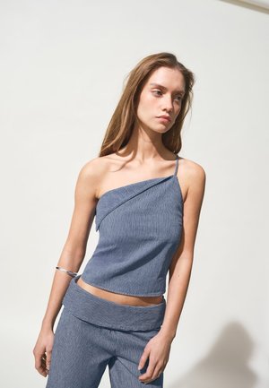 Mlada ženska z dolgimi lasmi, oblečena v modro črtast enoramni zgornji del in ujemajoče hlače, stoji pred enostavnim svetlim ozadjem.