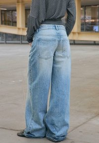 Persoană purtând blugi largi, colorați în albastru deschis și un tricou cu mânecă lungă, cu dungi negre și gri, stând într-un spațiu interior din beton.