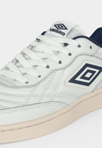 Λευκό αθλητικό παπούτσι Umbro με ναυτικό μπλε λογότυπο, λευκά κορδόνια και μπεζ σόλα, που δείχνει λεπτομερείς ραφές και πλέγμα στην γλώσσα.