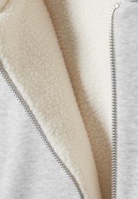 Nærbillede af cremefarvet fleeceforing indeni en lysegrå jakke eller sweatshirt med lynlås, der fremhæver tekstur og metallynlåsens tænder.