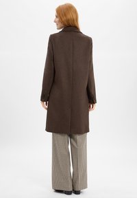Cappotto di lana marrone con una vestibilità su misura, colletto a punte e polsini abbottonati, abbinato a pantaloni a gamba larga a quadri. Texture uniforme, design minimalista.