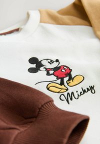 Mikki Hiire sweatshirt, millel on valge keha ja pruunid ning beežid varrukad. Kätkeb tikandiga graafikat Mikist ja kollaseid aktsente kingadel.