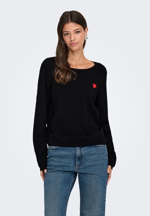 Mujer con suéter negro con un pequeño corazón rojo en el pecho y vaqueros azules, de pie frente a un fondo claro y liso.