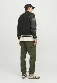 Chaqueta bomber negra con mangas de cuero, puños a rayas, combinada con pantalones cargo en color oliva y zapatillas beige. Vista trasera del atuendo.