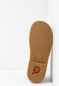 Semelle en caoutchouc avec une surface texturée, présentant un détail de logo rouge et noir. Le design est de couleur beige naturel avec une forme arrondie.