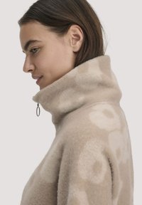 Vrouw met bruin haar draagt een beige, patroonfleecejack met een hoge ritssluiting, zijaanzicht tegen een effen achtergrond.