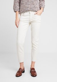 Jeans de mezclilla blanca con un corte ajustado, que cuentan con una cremallera en la parte frontal y bolsillos delanteros, combinados con zapatos de cuero marrón. Top con patrón texturizado.
