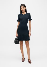 Γυναίκα που φορεί κολλητό μαύρο φόρεμα με κοντά μανίκια και βολάν, κρατώντας μαύρη τσάντα και φορώντας σκουροκόκκινα παπούτσια Mary Jane.