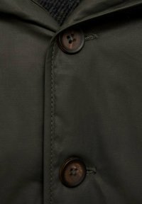 Tumman vihreä takki, jossa on lähikuva sileästä kankaasta, kaksi ruskeaa nappia, joissa on neljä reikää kummassakin, ja ommeltu yksityiskohta saumoissa.