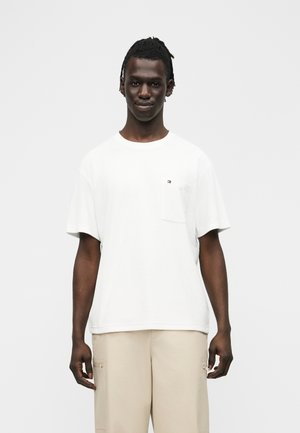 Junger Mann steht und trägt ein schlichtes weißes T-Shirt mit kleinem Logo auf der Brusttasche sowie beige Hosen vor einem einfarbigen Hintergrund.