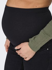 Bordură elastică neagră pentru maternitate combinată cu blugi închiși la culoare, mână odihnindu-se pe abdomen, bluză verde cu mâneci lungi și textură vizibilă.