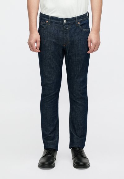 Jean en denim bleu foncé à coupe droite, avec cinq poches, fermeture par bouton en métal et détails de couture. Porté avec des chaussures noires.