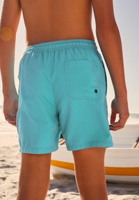 Türkiissinised ujumispüksid, millel on elastne vöökoht ja tagatasku, valmistatud kergetest materjalidest. Disain on lihtne, ilma mustriteta.