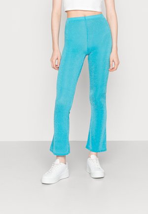 Calças flare turquesa com um acabamento texturizado, feitas de um tecido elástico. Combinadas com sapatilhas brancas para um look casual.