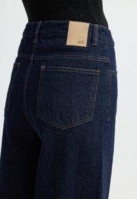 Ciemnoniebieskie dżinsy o wysokim stanie, z brązową skórzaną metką marki z tyłu oraz tradycyjnymi tylnymi kieszeniami z detalami przeszycia.