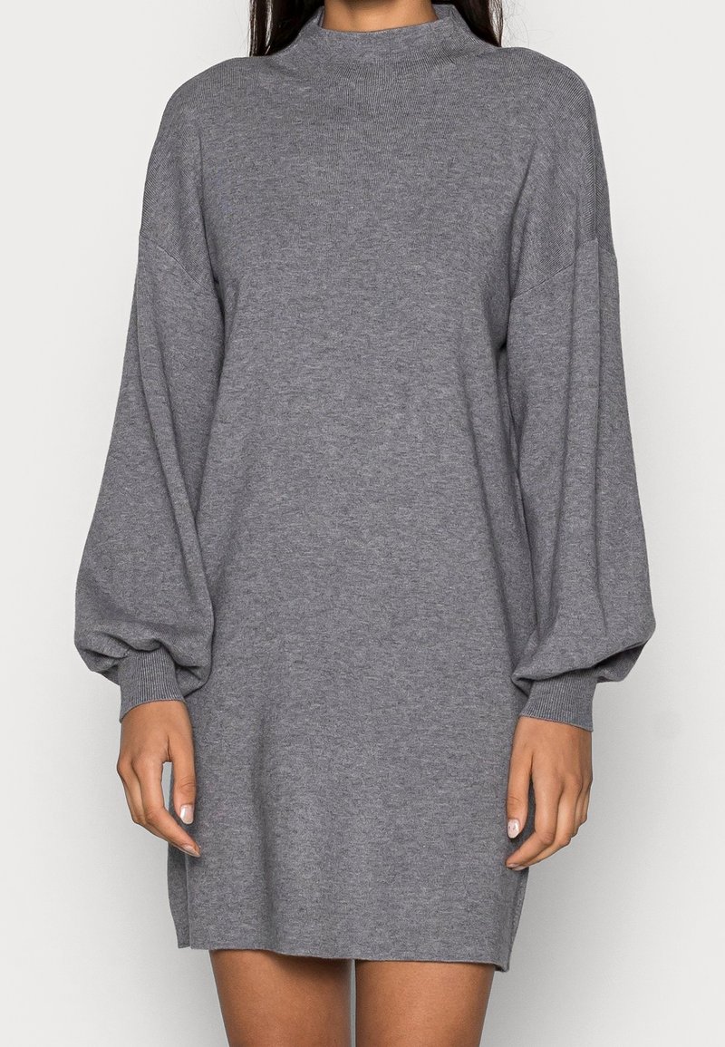 Femme portant une robe-pull en maille grise avec manches longues bouffantes et poignets côtelés, fond uni.