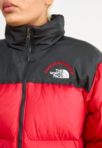 Rød og sort isoleret jakke med høj krave, der har The North Face-logoet og "Expedition System"-tekst i rød broderi.