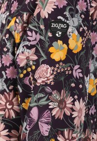 Bloemmotief stof met paarse, gele en roze bloemen, voorzien van een zwarte Zigzag logo patch aan de zijkant.
