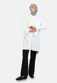 Camicia bianca a maniche lunghe con colletto e taschino sul petto, abbinata a pantaloni neri a righe sottili. La camicia presenta dettagli con cravatte sulle maniche.