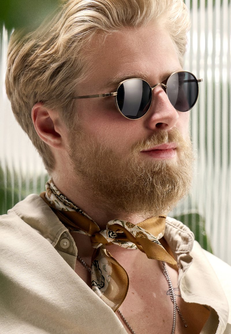 Pyöreät mustat aurinkolasit, joissa on hopeanvärinen metallikehys, yhdistettynä vaaleanruskeaan, kuvioituun silkkihuiviin, joka on solmittu kaulan ympäri, beige paidan yllä.