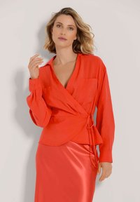 Rotes Wickelshirt aus leichtem Stoff mit langen Ärmeln, V-Ausschnitt und vorderem Bindedetail. Model trägt einen passenden roten Satinkleid.