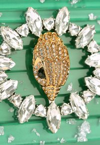 Spilla dorata a forma di conchiglia con strass chiari scintillanti, circondata da pietre a forma di navetta e a forma di cuore, su sfondo verde.