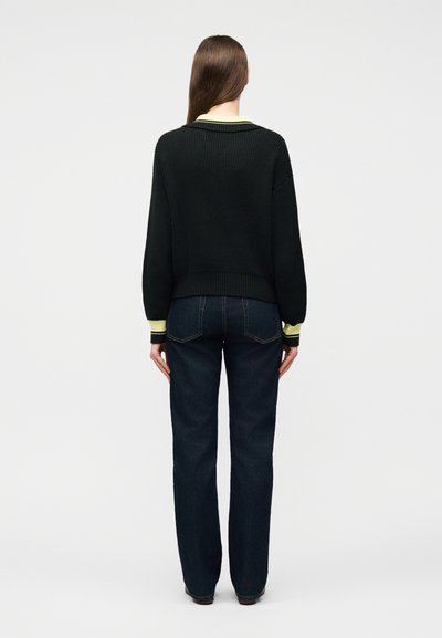 Γυναίκα με μακριά καστανά μαλλιά, φορούν μαύρο ριμπ πλεχτό πουλόβερ με κίτρινη λεπτομέρεια και σκούρο μπλε τζιν, στέκεται με την πλάτη προς τα εμπρός σε απλό φόντο.