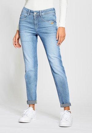 Frau trägt hellblaue, hochgekrempelte Jeans und weiße Turnschuhe, steht vor einem schlichten hellen Hintergrund.